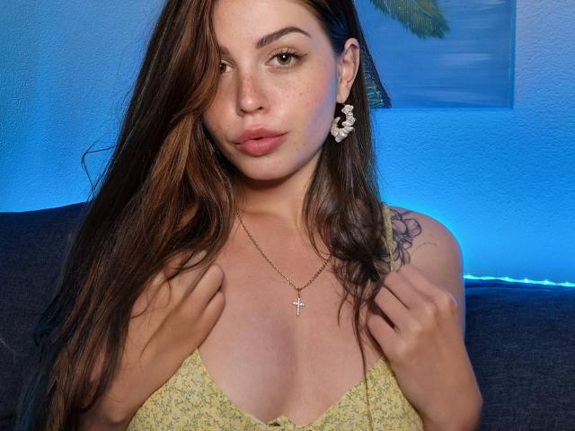 ValeriMair, avec de longs cheveux bruns, portant une robe jaune à motifs floraux, un collier en forme de croix et des boucles d'oreilles en forme de croissant de lune blancs, tenant les bretelles de la robe, montrant de petites seins et un tatouage sur son épaule droite..