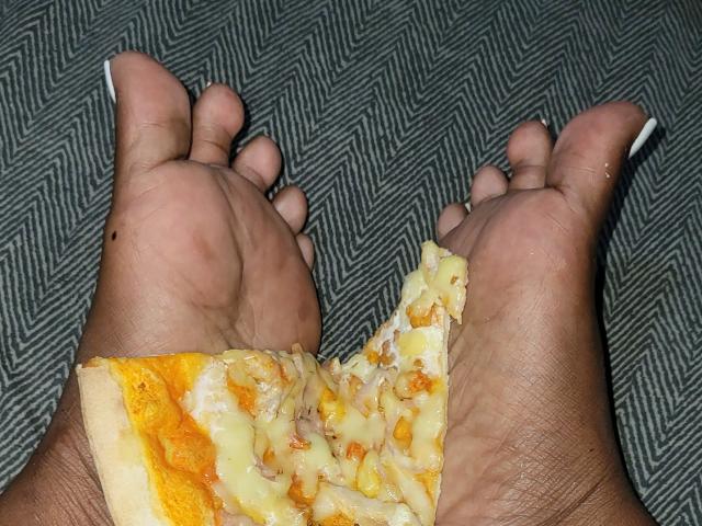 Los pies de ChristinneLov sostenían una porción de pizza, con queso y de color naranja, entre sus dedos. Sus uñas son blancas, y el patrón en el suelo es en zigzag..