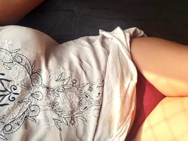 MeahBrunette's pale thighs and white dress with black floral embroidery, lying on a red and black bed. Sunlight casts soft shadows on her skin.