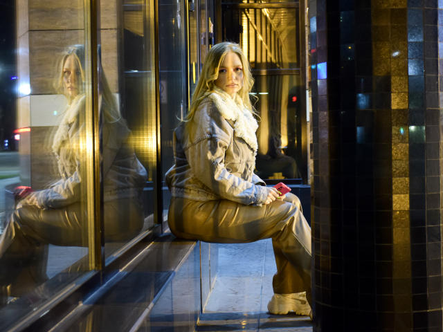 SweetAleksaX met lang blond haar, zittend op een stoel, gekleed in een glanzend gouden jas en broek, met een rode telefoon in haar hand, reflectie in glas, stedelijke omgeving 's nachts..