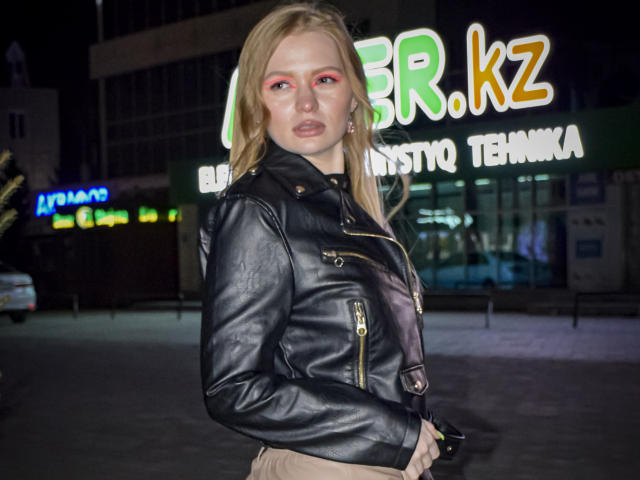 SweetAleksaX con capelli biondi, trucco rosso sugli occhi, giacca di pelle nera e pantaloni beige, in una foto notturna, che tiene un telefono, guardando direttamente la fotocamera..