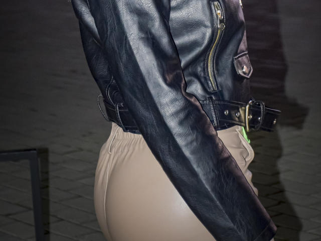 SweetAleksaX con una chaqueta de cuero negra, trasero desnudo en pantalones ajustados, cinturón negro con hebilla dorada, de pie con los brazos cruzados sobre la chaqueta, correa verde visible, noche, suelo de adoquines..