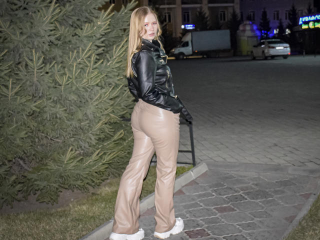 SweetAleksaX, com cabelos loiros longos, jaqueta de couro preta e calças de couro bege, em pé em um caminho de tijolos, olhando por cima do ombro, mostrando suas nádegas, usando botas brancas..