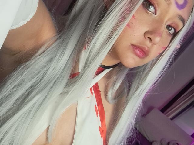 KuruChan, con lunghi capelli bianchi, un segno viola sulla fronte, trucco rosso e labbra rosa, indossa una maglietta bianca con spalline rosse, ha un seno piccolo e un viso carino..