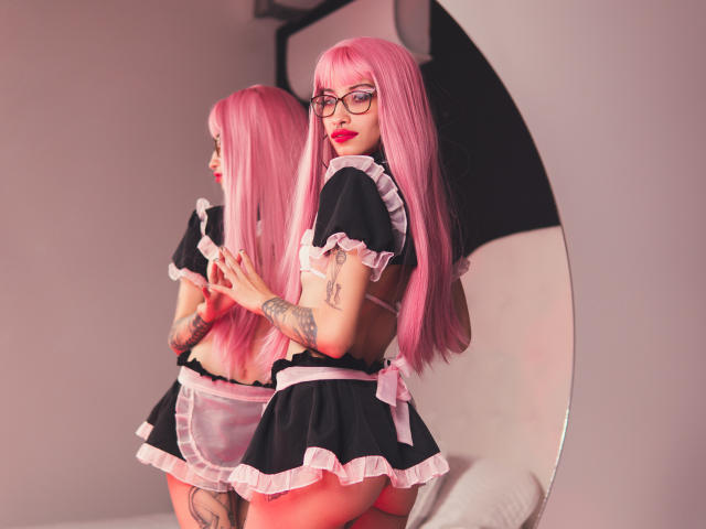 Laila Stewart, con i capelli rosa, gli occhiali e un abito da cameriera bianco e nero, mostra la schiena e i tatuaggi, guardando oltre la spalla, in piedi di fronte a uno specchio..