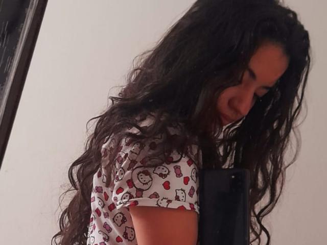 NicolSky with long, wavy black hair, wearing a white shirt with red and black cat prints, holding a black phone, looking down with a subtle smile.