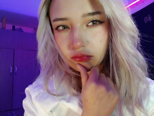 KuruChan ha i capelli biondo platino, labbra rosa carnose e sta toccando il mento con un dito. Indossa una camicia bianca, con una leggera illuminazione rosa e viola..
