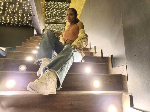 CinthiaWalker, with medium tits, sits on wooden stairs, wearing a pink crop top, blue jeans, and white sneakers, under warm lights, looking up with a slight smile.