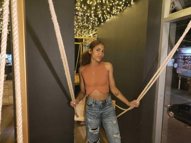 CinthiaWalker with a slim, toned body, wearing a cut-out, pink, sleeveless top and ripped jeans, holding thick ropes, standing in a dimly lit, string-lit room.