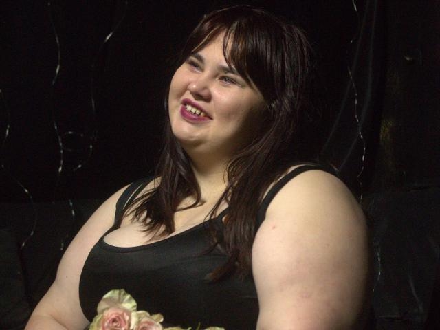 KaidoKun, una mujer robusta con cabello castaño oscuro, sonriendo con lápiz labial rosa, vestida con una camiseta negra, mostrando sus senos, sosteniendo un ramo de rosas..