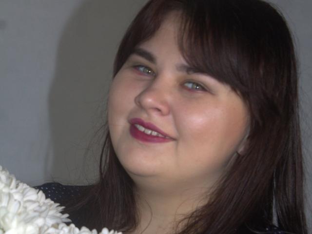 KaidoKun a un visage mignon et rond, avec des cheveux brun foncé, une peau claire et du rouge à lèvres rose. Elle sourit, et un objet blanc et duveteux se trouve au premier plan..