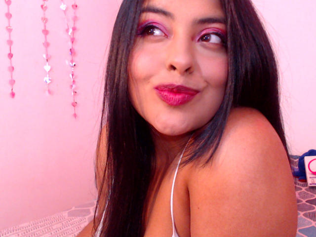 AngelaRoggers, com sombra rosa, batom vermelho e cabelo preto, sorrindo para a câmera, mostrando o ombro nu e seios pequenos, vestindo uma blusa branca de alças finas..
