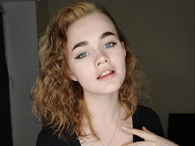 Anna, com cabelo ruivo cacheado, pele clara, olhos azuis, delineador preto e um leve sorriso, tocando o peito com a mão, vestindo uma blusa preta..