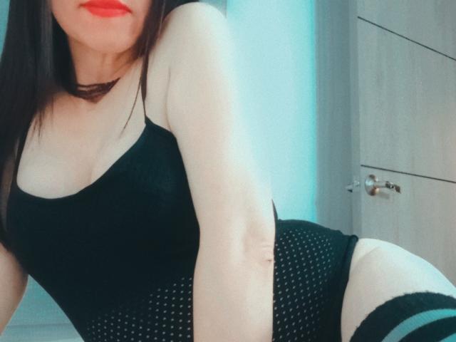 MondBlue ha la pelle chiara, labbra rosse, biancheria intima nera a pois e calze nere che arrivano sopra le cosce, è china in avanti mostrando i glutei e piccoli seni, con un braccio alzato per un selfie..