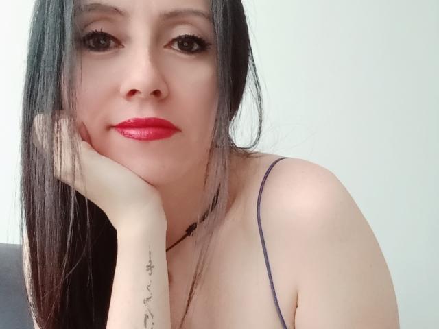 MondBlue has long black hair, red lipstick, and a tattoo on her right wrist. She's resting her chin on her hand, wearing a purple spaghetti strap top.