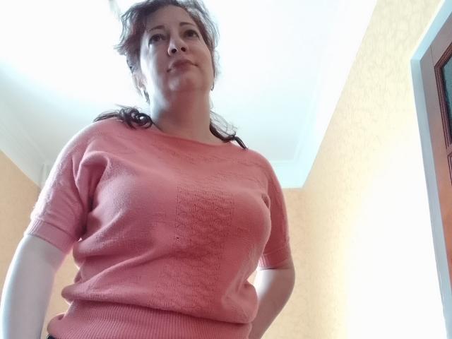 LediSusana, een mollige vrouw met bruin haar, draagt een roze, gestructureerde trui en staat met haar handen op haar heupen, kijkend naar de camera met een lichte glimlach..