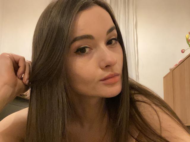 Dionisia with long brown hair, slightly parted lips, and dark eyebrows, looking to the side, her hand in her hair, showing off her smooth, pale-skinned face and subtle makeup.