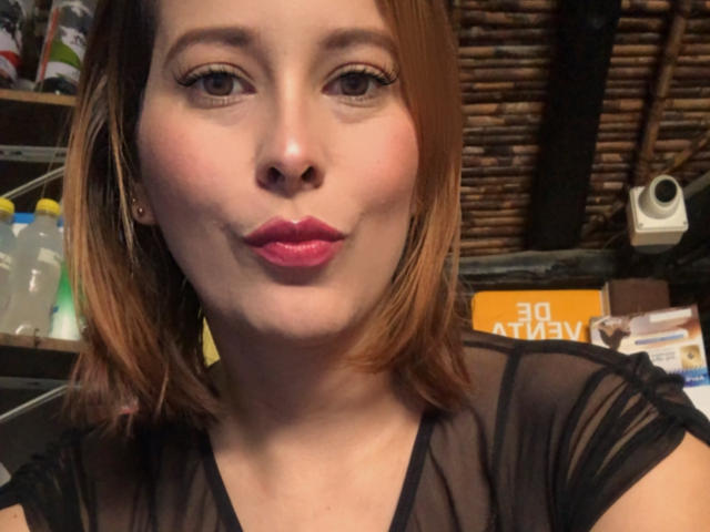 Sarita Velez, con rossetto rosa, occhi marroni e capelli castani di lunghezza media, indossa una maglia nera trasparente, fa un'espressione languida e mostra il suo viso carino e sensuale..