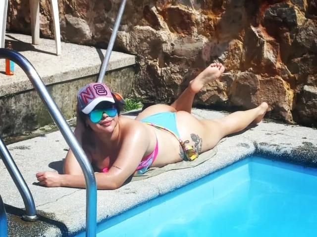 EveNailah liegt am Pool, trägt einen pink-blauen Bikini, eine weiße Kappe und eine Sonnenbrille, hat das Gesäß hoch und die Beine angehoben und zeigt dabei ein Tattoo..