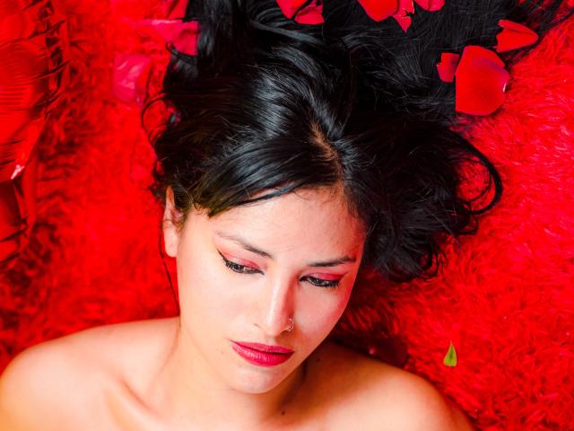 SilvannaReyes with dark hair, red lipstick, and pink eyeshadow, lying on red fur, surrounded by red rose petals, looking down with a slight frown, nose pierced.