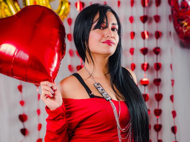 Silvanna Reyes, con cabello negro largo, vestido rojo con un hombro descubierto, sosteniendo un globo rojo brillante con forma de corazón, con una correa de sostén negra y un collar de plata, sonriendo a la cámara..
