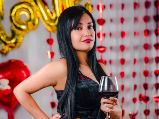 SilvannaReyes with long black hair, red lipstick, black velvet dress, holding a glass of red wine, standing, smiling, golden balloons, red heart decorations in the background.