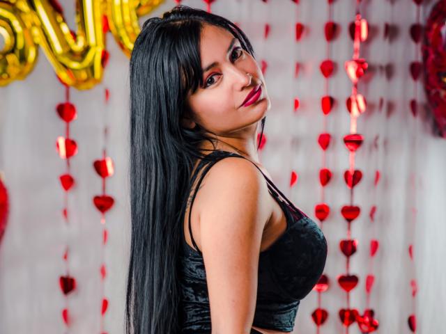 Silvanna Reyes mit langem, schwarzem Haar, die über ihre Schulter schaut, trägt ein schwarzes Samtkleid, das ihre Brüste zeigt, rote Lippenstift und einen Nasenring, in einer festlichen Umgebung..