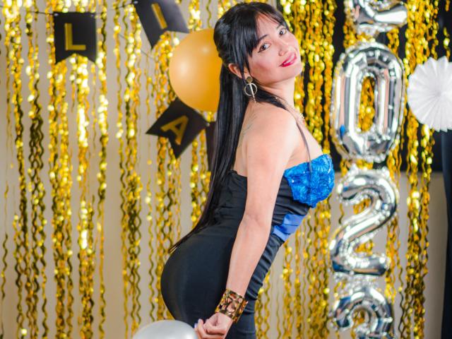 Silvanna Reyes, con lunghi capelli neri, rossetto rosso, abito nero e blu senza spalline che mette in mostra il sedere, un braccialetto dorato, un palloncino arancione, un palloncino argentato con il numero "20" e festoni dorati sullo sfondo..