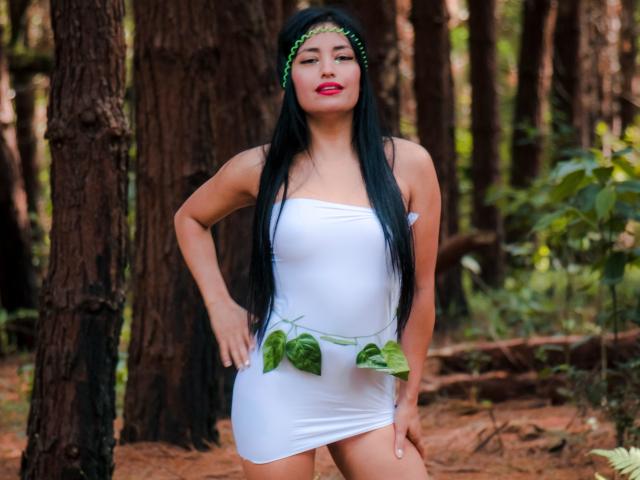 SilvannaReyes has long black hair, red lipstick, and a green leaf headband. She's wearing a tight white dress with green leaves around her waist, showing her small tits.