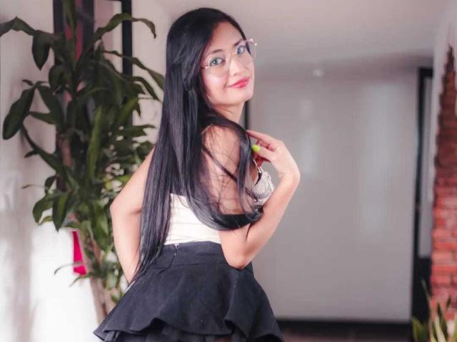 Silvanna Reyes, met lang zwart haar, blauwe ogen en een bril, lacht naar de camera, draagt een witte top en een zwarte rok, en streelt haar haar, terwijl ze in een kamer staat..