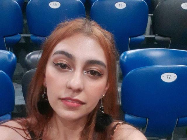 Rosalie Black hat rotes Haar, braune Augen mit Lidstrich und trägt kleine Ohrringe. Sie sitzt auf blauen Stadionstühlen, auf denen "18" und "19" stehen..