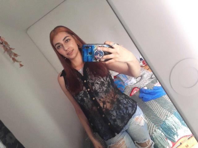 RosalieBlack, avec de longs cheveux roux, portant une chemise noire en dentelle sans manches et un jean déchiré, prend un selfie devant un miroir avec un téléphone bleu, affichant un léger sourire..