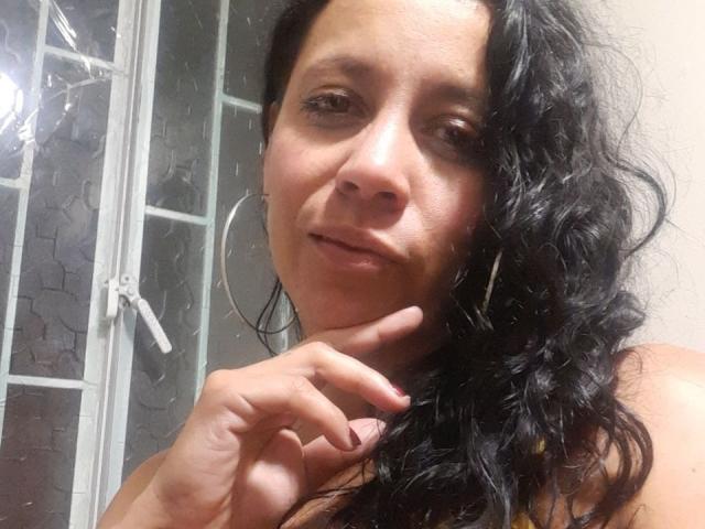 IsabellaSweett heeft krullend zwart haar, een medium bruine huidskleur en ze glimlacht lichtjes met haar vinger tegen haar kin. Ze draagt grote oorringen en heeft een subtiele, natuurlijke uitstraling..