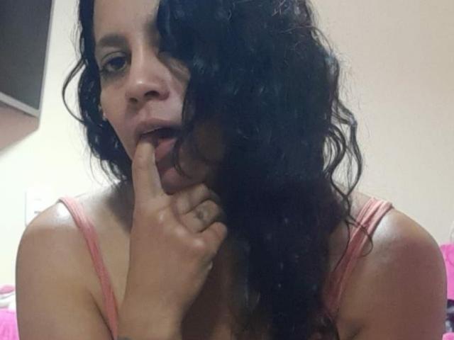 IsabellaSweett mit lockigem, schwarzem Haar, rosa Tanktop, Finger im Mund, leicht geöffneten Lippen, gebräunter Haut, konzentriertem Gesichtsausdruck, mittelgroßer Brust, weichem Licht im Schlafzimmer..
