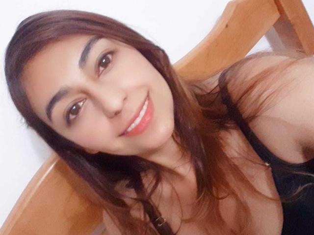 RosalieBlack, met bruin haar, lachend, gekleed in een zwarte top, waarbij haar borsten zichtbaar zijn, zittend op een houten stoel, witte achtergrond, selfie, rode lipstick, bruine ogen, medium huidskleur..