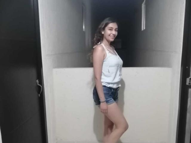 RosalieBlack, avec ses cheveux bruns ondulés, son débardeur blanc et son short en jean, se tient dans un couloir, souriante, montrant sa petite poitrine et ses jolies fesses, et portant un bracelet noir..