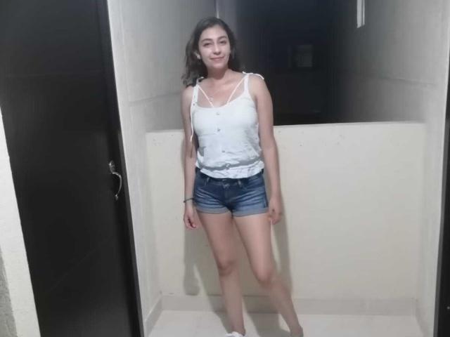 RosalieBlack with medium tits in a white tank top and denim shorts, standing in a narrow room, smiling with brown hair, and wearing white flats.
