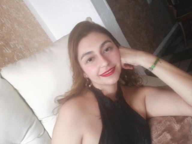 RosalieBlack, avec ses cheveux bruns ondulés, son rouge à lèvres rouge et son haut noir à encolure dégagée, sourit à l'appareil photo, sa main reposant sur sa joue, un bracelet vert à son poignet..