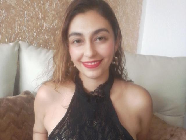 RosalieBlack met golvend bruin haar, rode lippenstift, lachend, gekleed in een zwart kanten topje met halter, waarbij haar kleine borsten en blote schouders zichtbaar zijn, zittend op een beige bank..