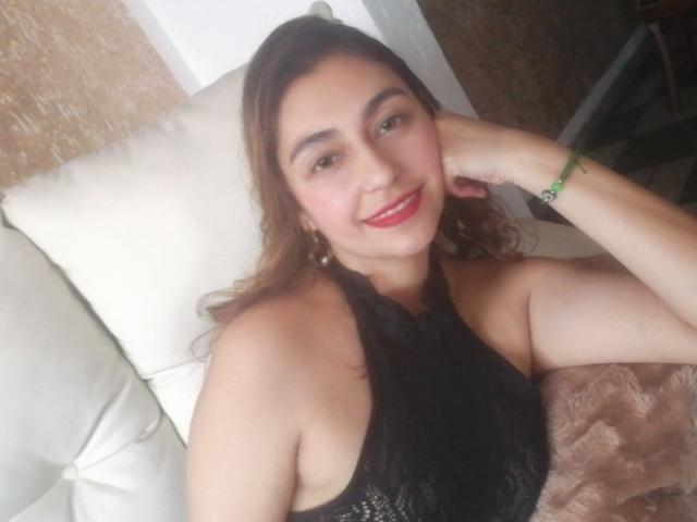 RosalieBlack with red lipstick, smiling, black halter top, showing her tits, lying on a white pillow, brown hair, green bracelet, hand on cheek, cute, bedroom.