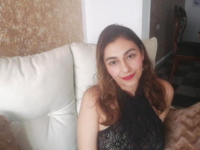 RosalieBlack, con cabello castaño ondulado, lápiz labial rojo y una blusa de encaje negro, sonriendo a la cámara, sentada en un sofá blanco, mostrando sutilmente sus pechos a través del encaje..