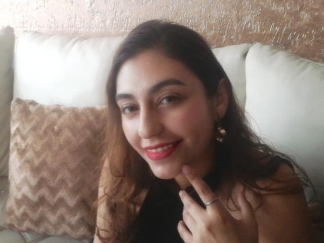 RosalieBlack, com cabelo castanho ondulado, batom vermelho, sorrindo, tocando o queixo com um dedo com anel, vestindo uma blusa preta, sentada em um sofá branco com uma almofada bege em padrão de chevron..