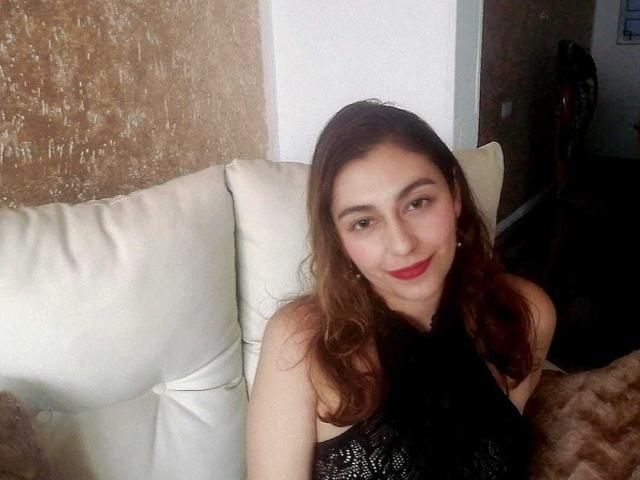 RosalieBlack tem batom vermelho, cabelo castanho ondulado e uma blusa preta de renda, está sentada em um sofá branco e sorrindo. Suas nádegas não estão visíveis..