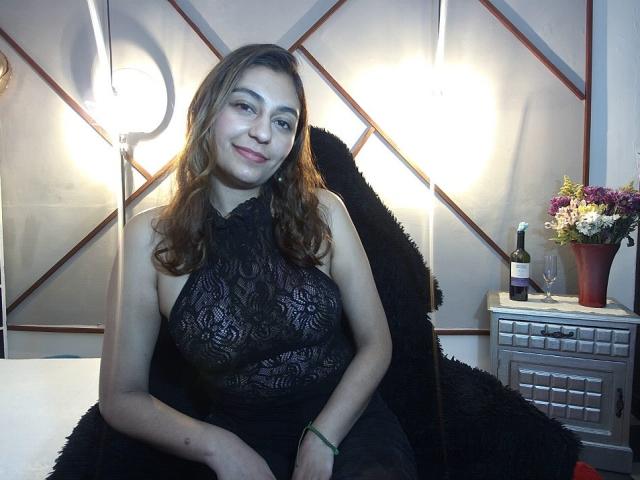 RosalieBlack, con lunghi capelli castani, sorridente, indossa un abito nero in pizzo con spalline, che mette in mostra il suo seno prosperoso, seduta su una poltrona nera e pelosa, con una bottiglia di vino e dei fiori..