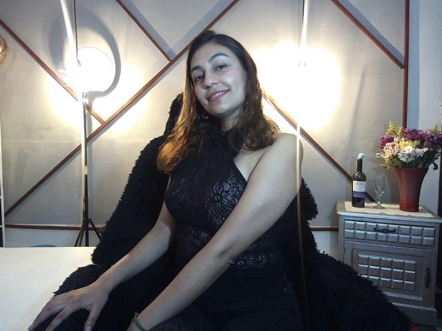 RosalieBlack, con cabello castaño, vestido de encaje negro y abrigo de piel negra, sentada, sonriendo, con luz en el fondo, una botella de vino, un vaso, flores en un mueble de madera, a la derecha..