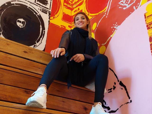 Stephaniez with tits covered, wearing a black scarf, black pants, and white sneakers, sitting on wooden stairs, smiling, blonde hair, black bag, red and yellow wall.