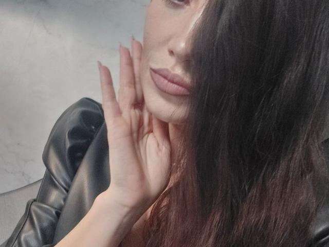 StassiLyanne ha la mano vicino al viso, con lunghi capelli castani che coprono un occhio. Indossa una giacca di pelle nera e le sue labbra sono leggermente tumefatte..