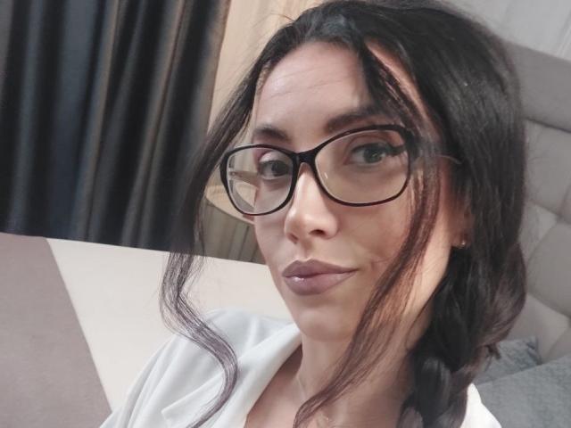 StassiLyanne, con i capelli scuri e ondulati, indossa occhiali neri, una camicia bianca e un trucco leggero, mostrando un'espressione sicura, con un leggero sorriso. Le sue natiche non sono visibili..