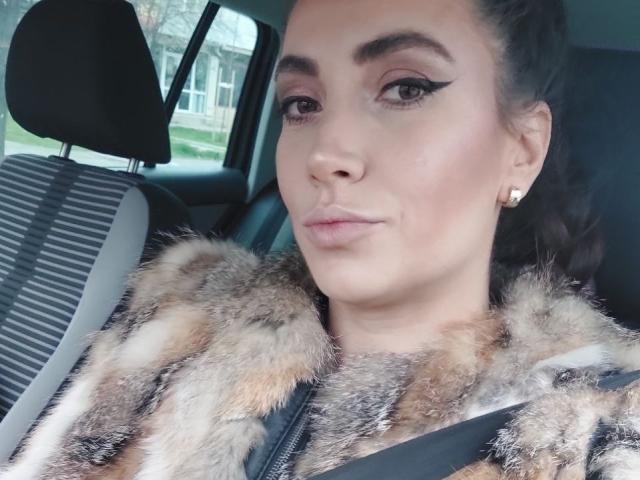 StassiLyanne mit einem flauschigen, braunen Fellmantel, schwarzem Eyeliner und Ohrringen, die eine verzogene Miene in einem Auto zeigt, Sicherheitsgurt angelegt, dunkles Haar im Pferdeschwanz..