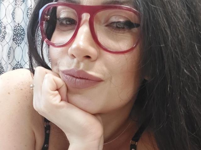 StassiLyanne, con gafas rojas, cabello negro y labios rosados, apoyando el mentón en la mano, mostrando su linda nariz y sus sutiles pecas. Está mirando a la cámara..